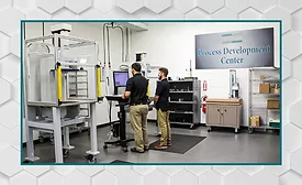 Engineers operating a materials testing system in a process development center, using automated testing equipment to evaluate product performance and manufacturing processes.